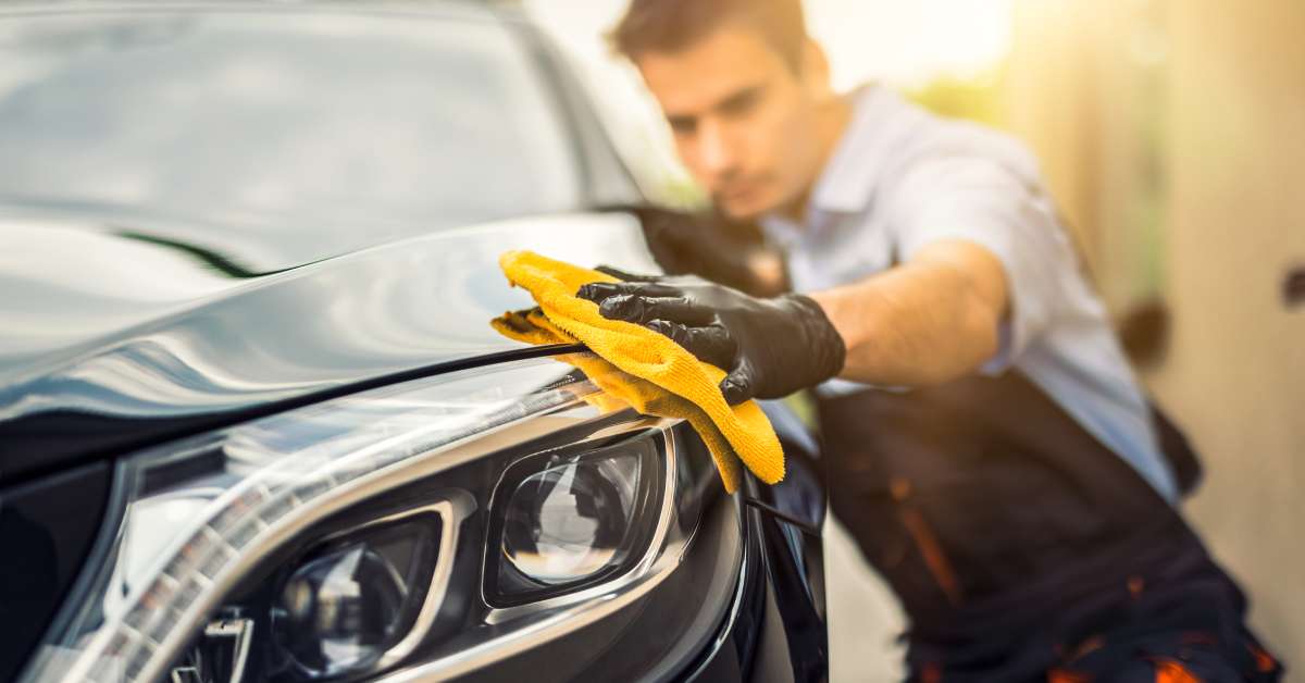 A professional detailer wearing overalls and gloves uses a cloth to polish the surface of a black car.